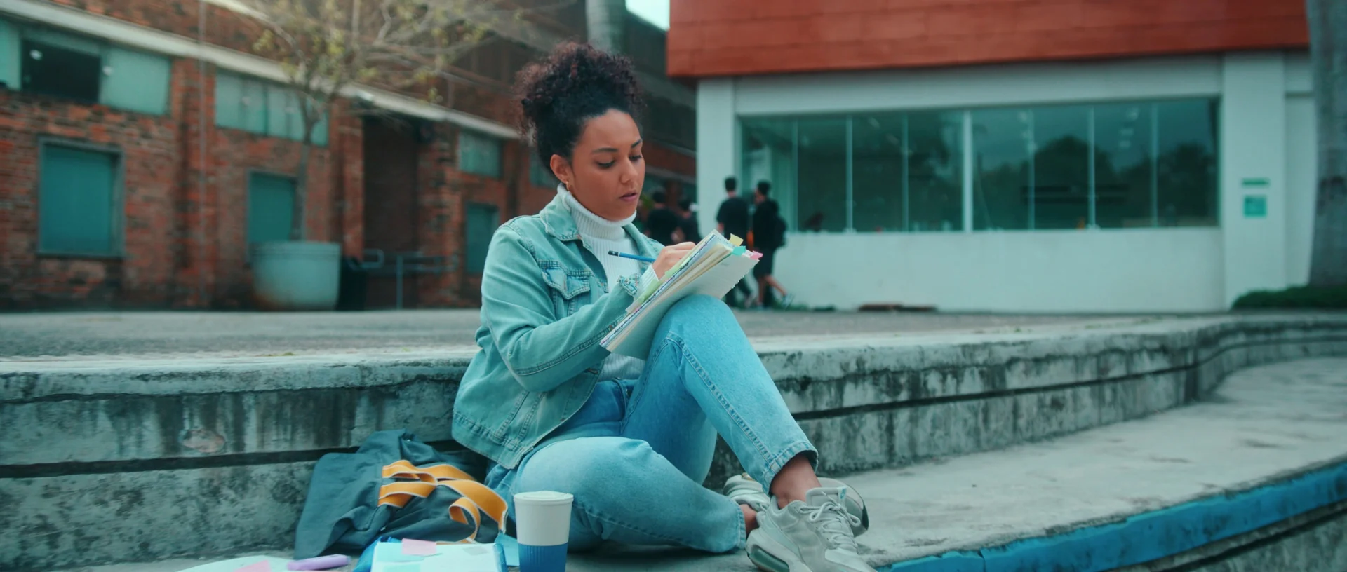 Jovem de jaqueta jeans escreve num caderno sentada nos degraus de um pátio externo, com mochila e copo ao lado e prédios de tijolo ao fundo.