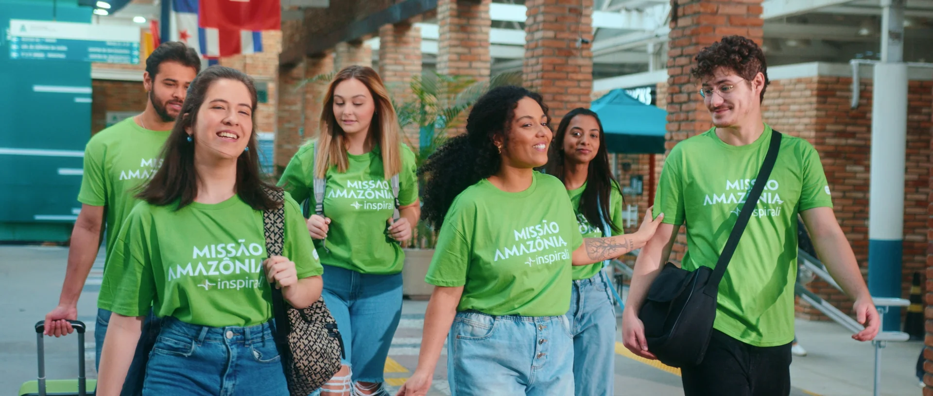 Grupo de estudantes sorridentes de camiseta verde da Missão Amazônia caminha com malas por um pátio de tijolos aparentes.