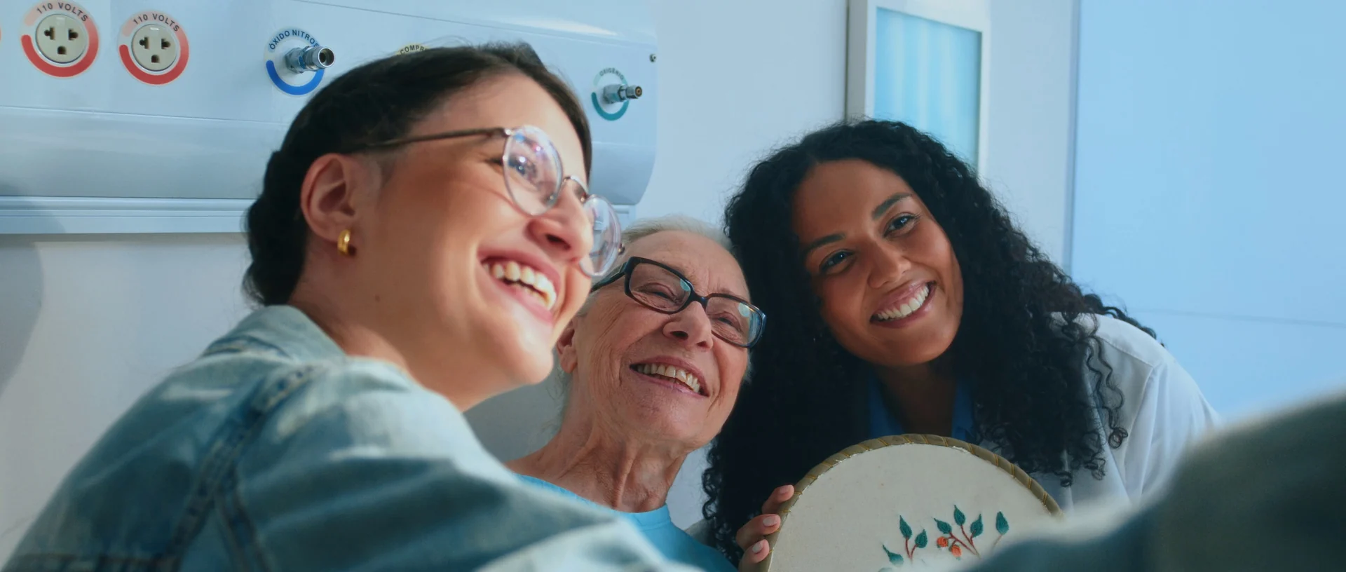 Médica de jaleco, uma mulher de jaqueta jeans e uma paciente idosa de óculos sorriem juntas num quarto de hospital, segurando um bordado.