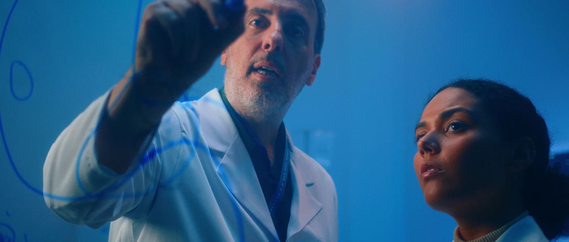 Professor de jaleco desenha com caneta num vidro, em luz azul, enquanto uma estudante ao lado acompanha com atenção.