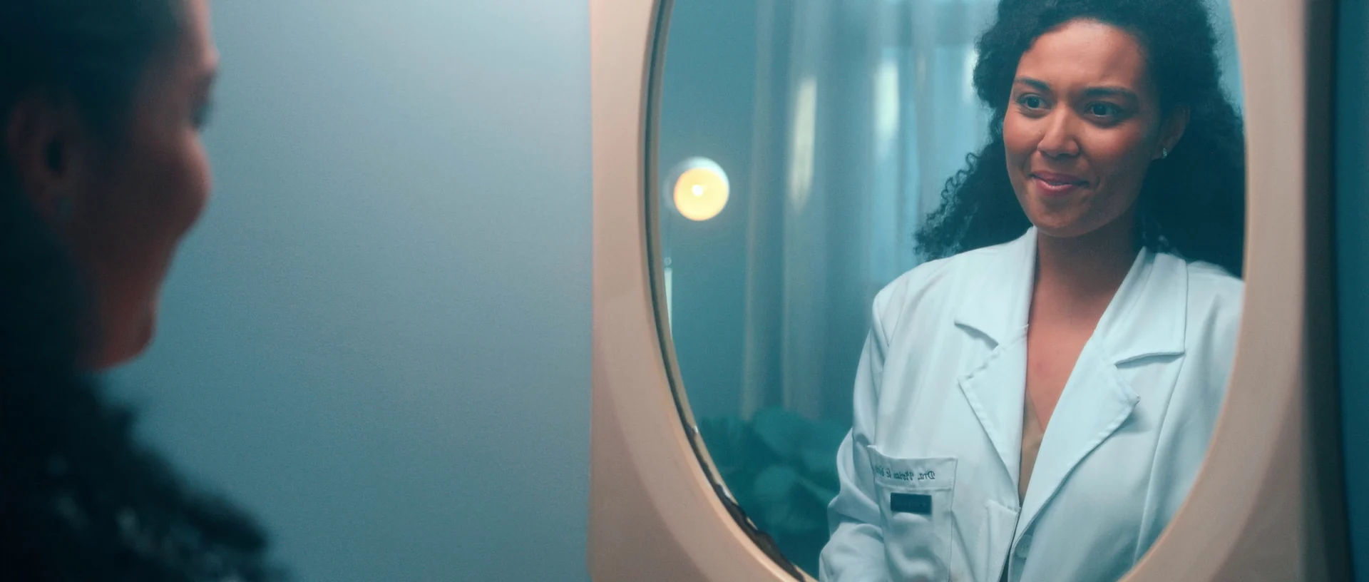Médica de jaleco branco sorri ao se ver num espelho redondo, num ambiente de luz suave.