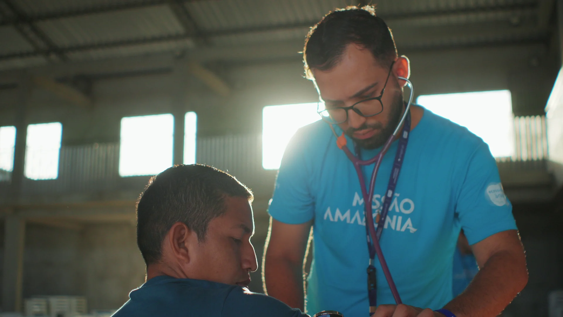 Estudante de medicina de camiseta Missão Amazônia ausculta um homem com estetoscópio, com luz de janela ao fundo.