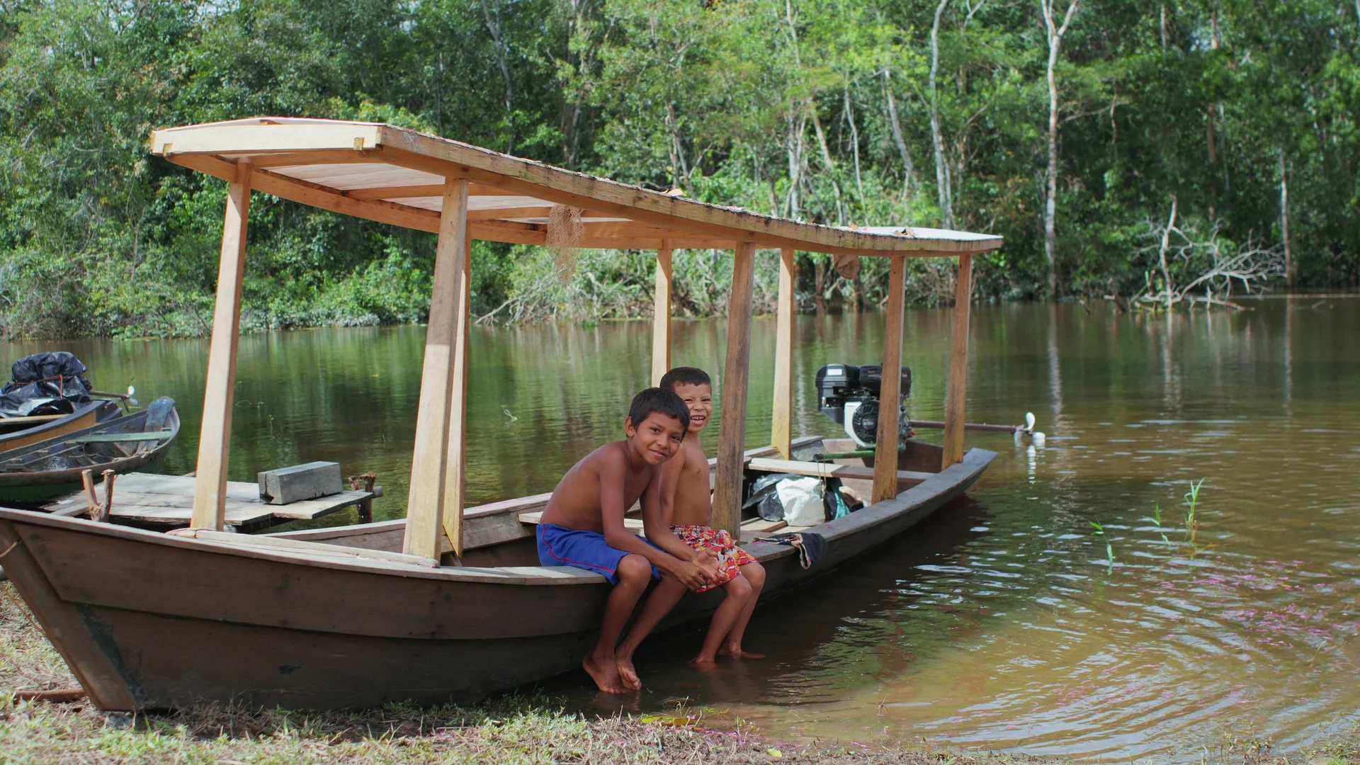 Dois meninos sentados na proa de uma canoa coberta, parada na água junto à mata.