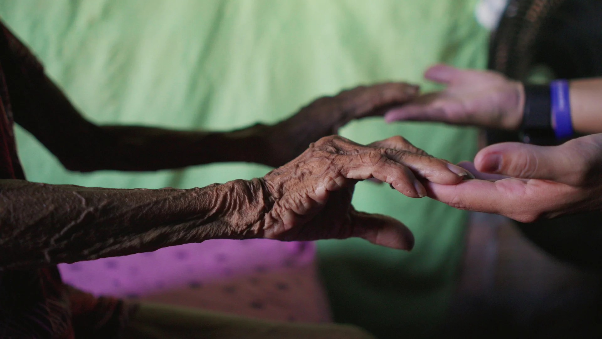 Mãos de uma pessoa idosa, marcadas pelo tempo, tocam as mãos de uma estudante.