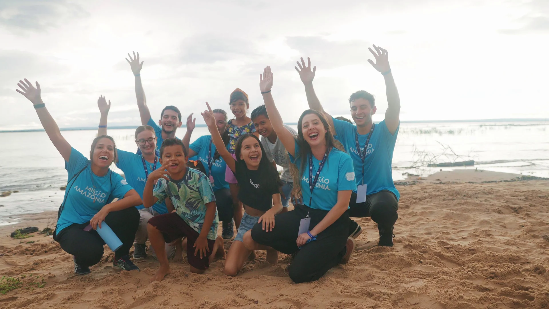 Estudantes da missão e crianças da comunidade comemoram com os braços para cima em uma praia de rio.