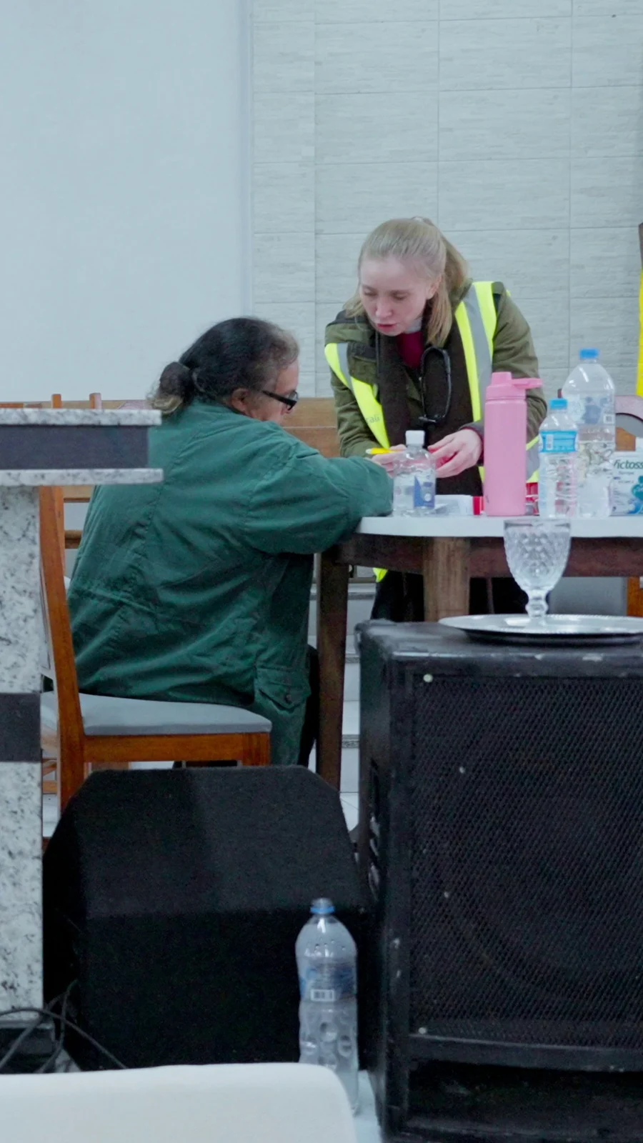 Voluntária de colete refletivo atende uma mulher sentada a uma mesa improvisada no abrigo.