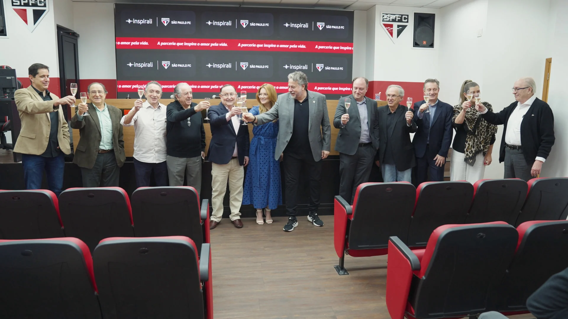 Grupo de pé diante do painel da parceria, no auditório do clube, ergue taças de espumante em brinde.