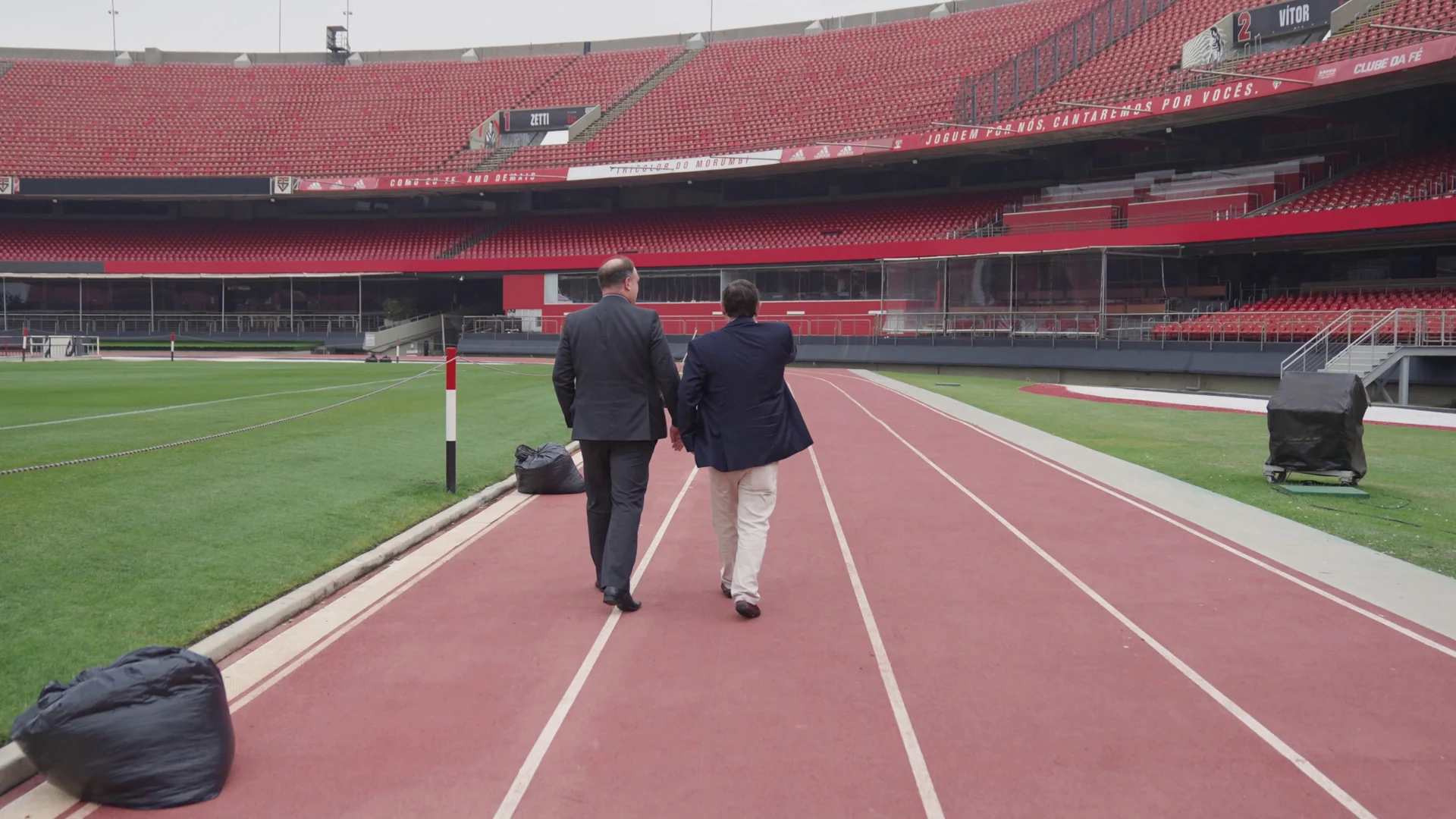 Dois homens de terno caminham de costas pela pista de atletismo ao lado do gramado do Morumbi.