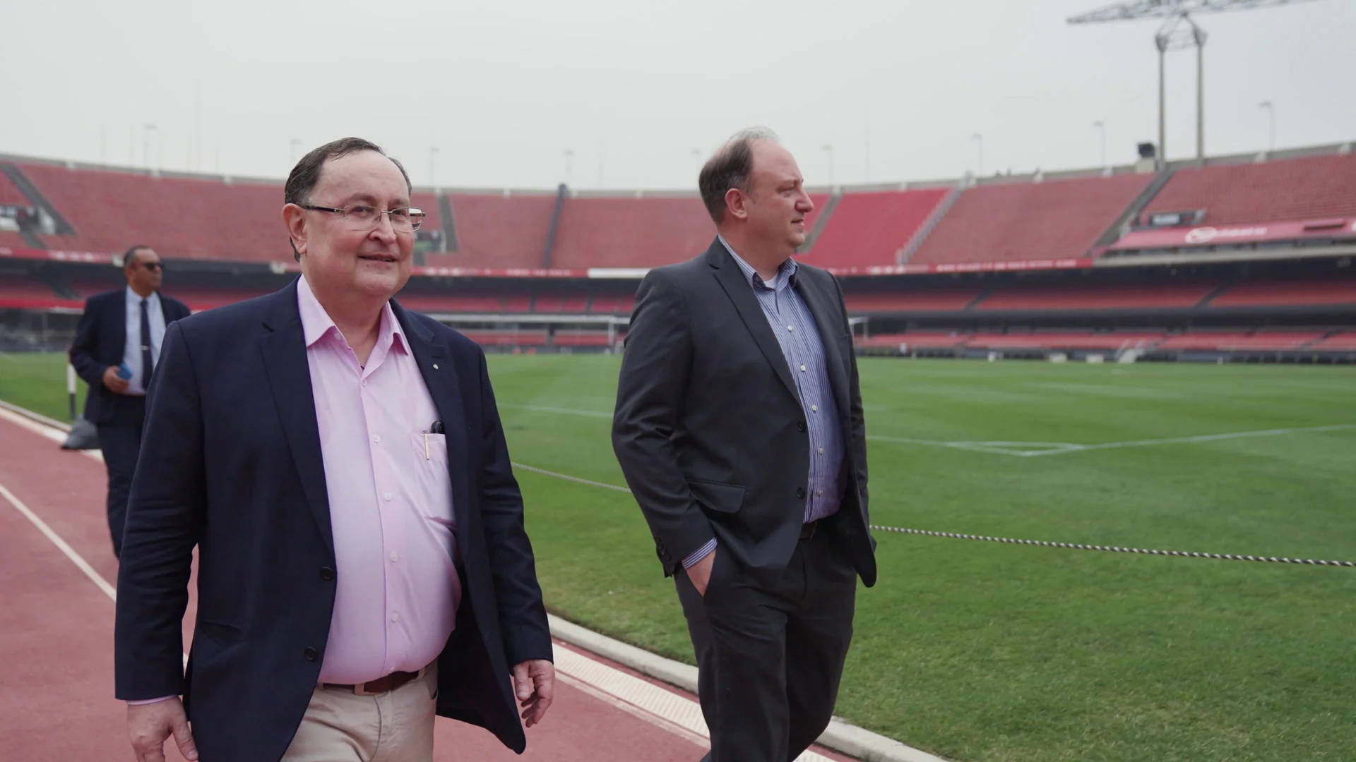 Dois homens de terno conversam em pé no gramado, com as arquibancadas vermelhas ao fundo.