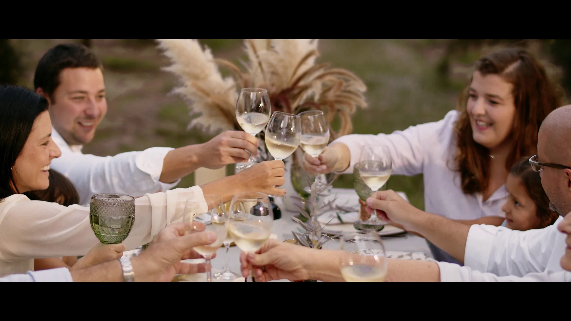 Família brindando com taças em volta de uma mesa posta ao ar livre, com capim-dos-pampas ao centro