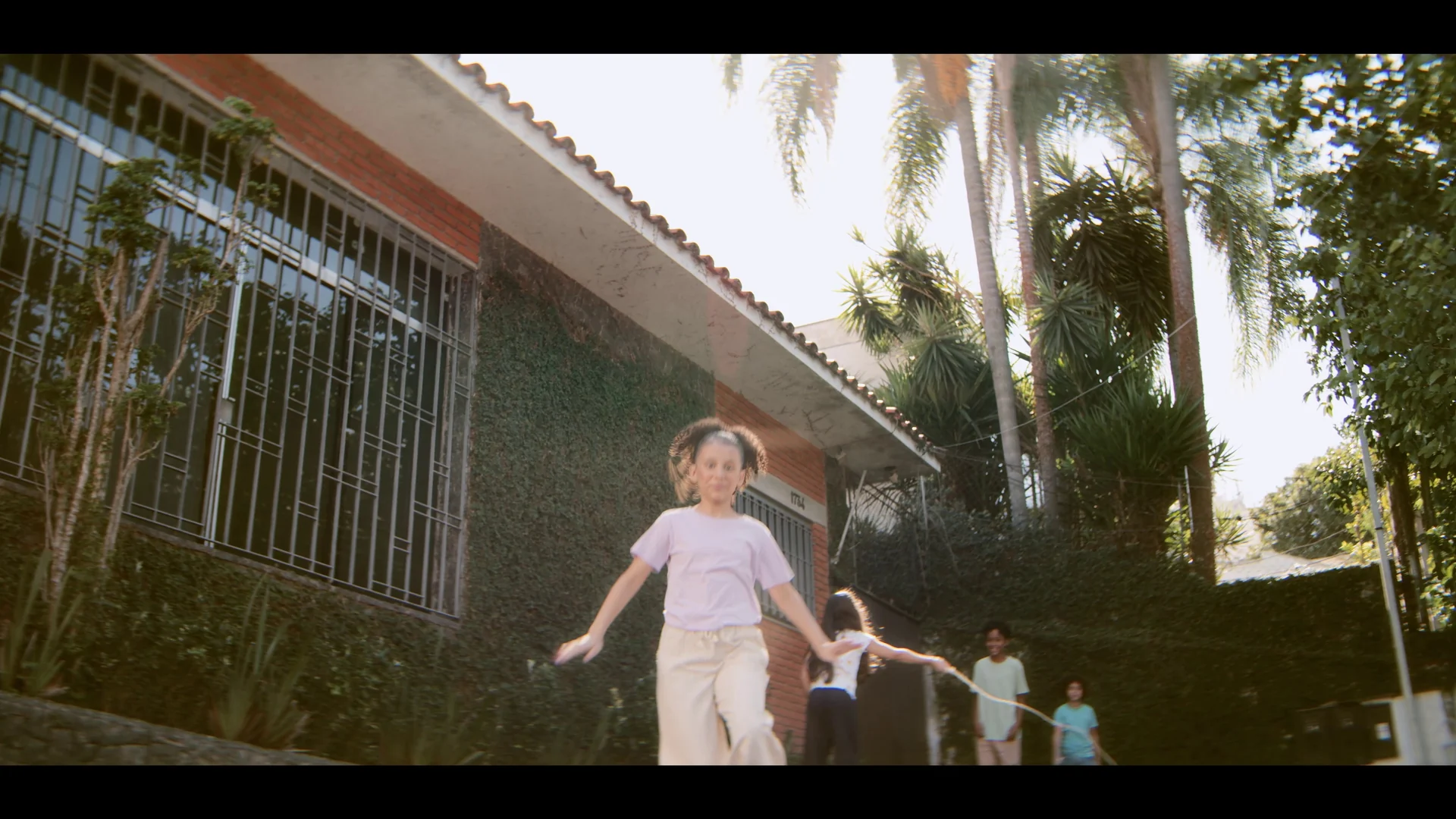 Menina de camiseta lilás corre sorrindo pelo jardim de uma casa de tijolos com palmeiras, em contraluz de fim de tarde.