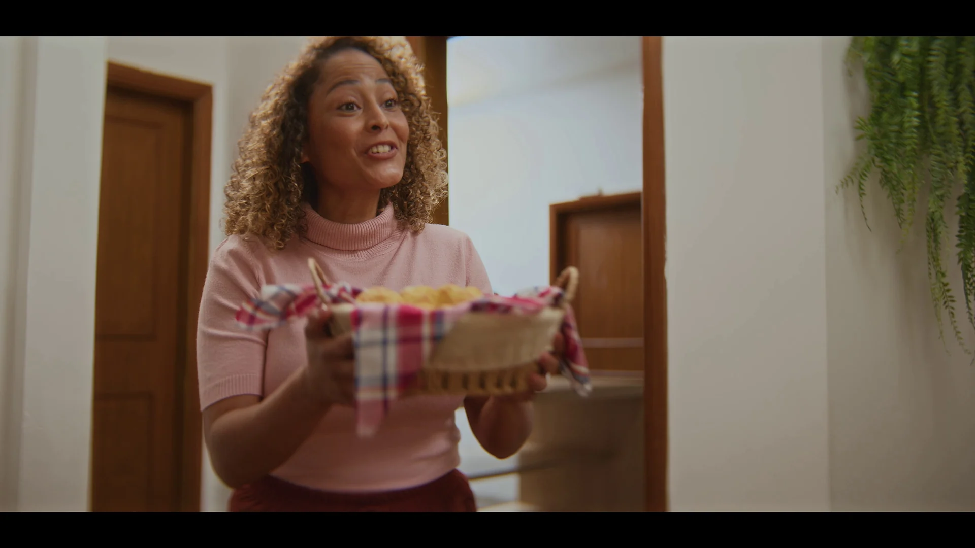 Mulher de cabelo cacheado e blusa rosa carrega uma cesta com pão de queijo forrada de pano xadrez, num corredor com portas de madeira.