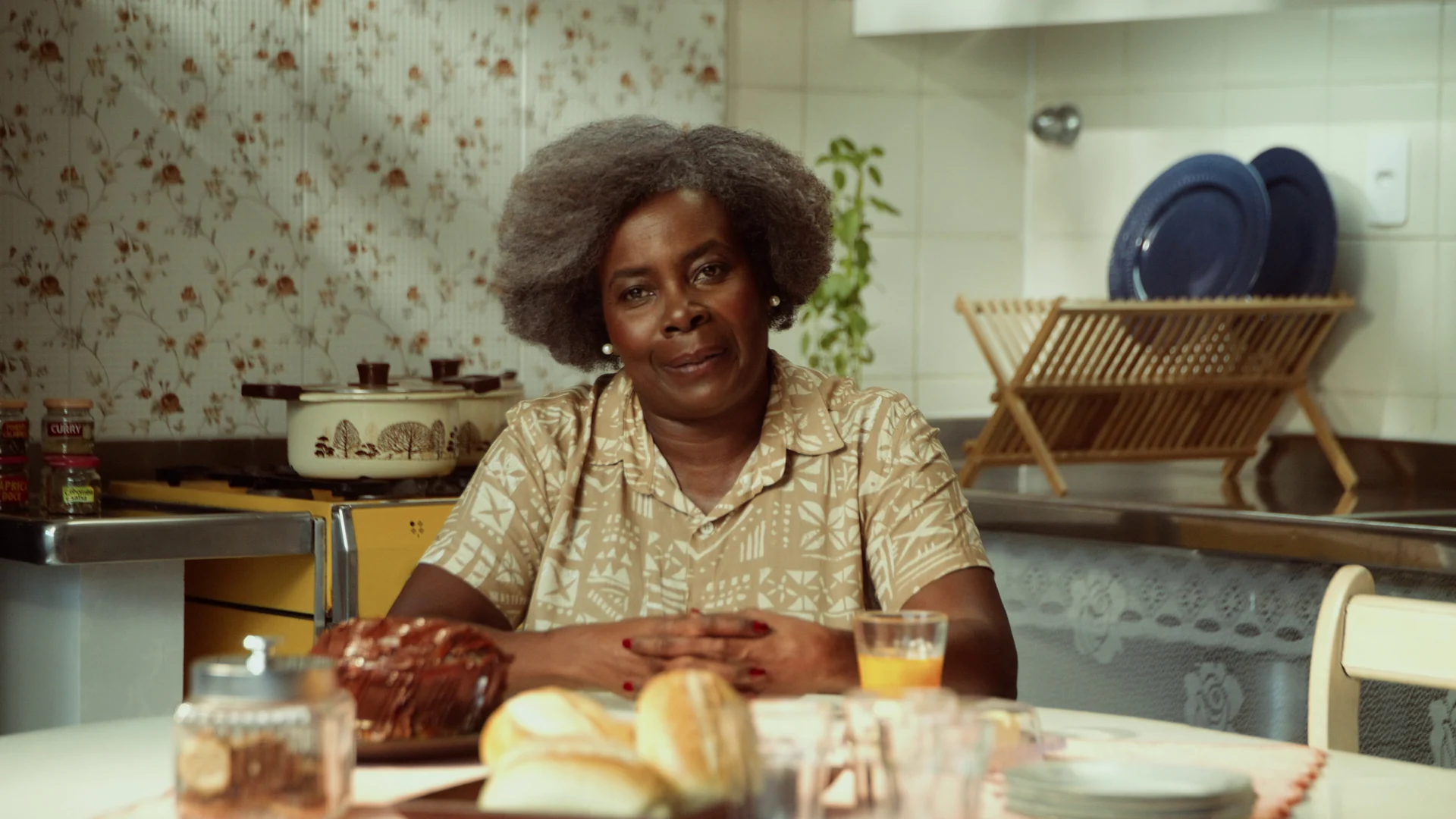 Senhora de cabelo grisalho sorri sentada à mesa de uma cozinha com papel de parede floral, com pães, bolo e suco diante dela.