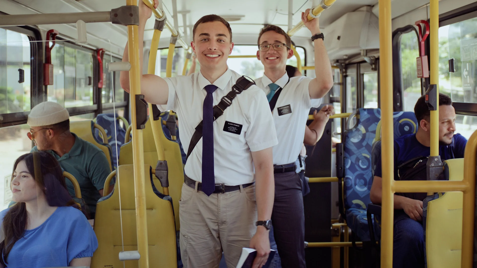 Dois missionários de camisa branca seguram nas barras amarelas de um ônibus urbano e sorriem para a câmera, com passageiros ao fundo.