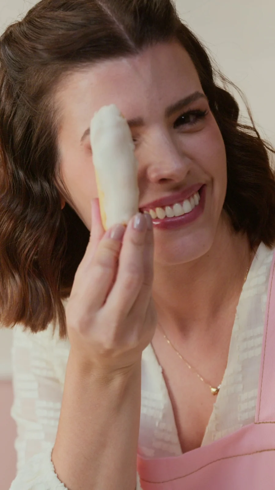 Ju Ferraz, de avental rosa, sorri segurando uma éclair de glacê branco perto do rosto.