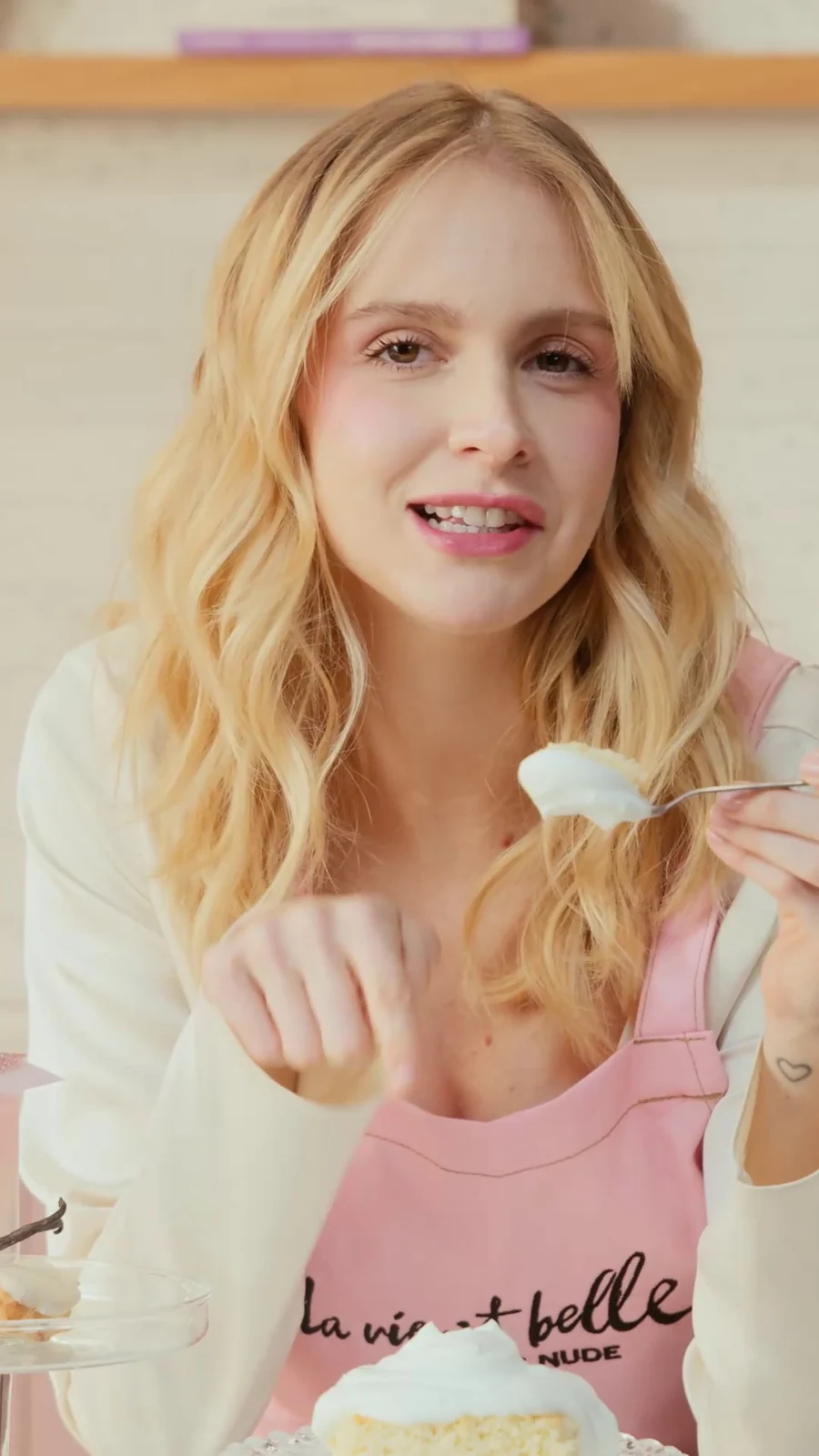 Isabella Scherer, de avental rosa, fala com uma colher na mão diante de uma fatia de bolo com chantilly.