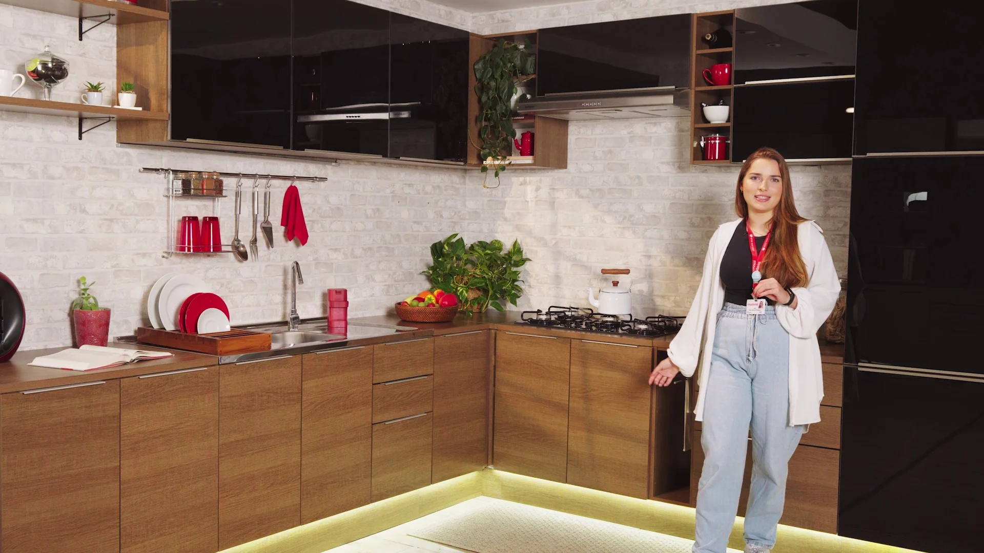 Apresentadora de jaleco branco em cozinha em L que combina balcões amadeirados e aéreos pretos, com luz de LED sob os módulos.