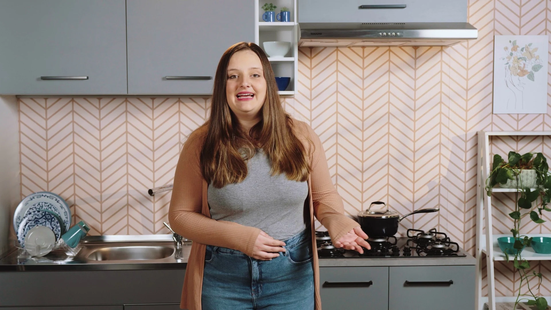 Apresentadora gesticula em cozinha com balcões cinza e rosa e revestimento em espinha de peixe rosado.
