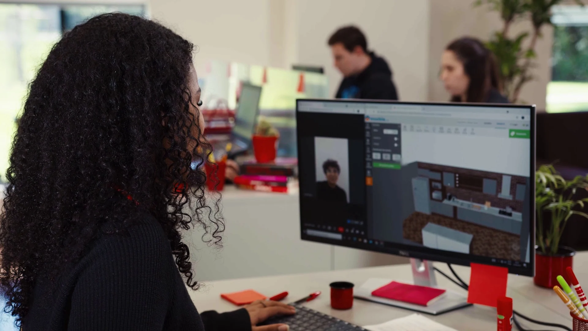 Projetista trabalha numa cozinha em software 3D durante videochamada, com colegas ao fundo no escritório.