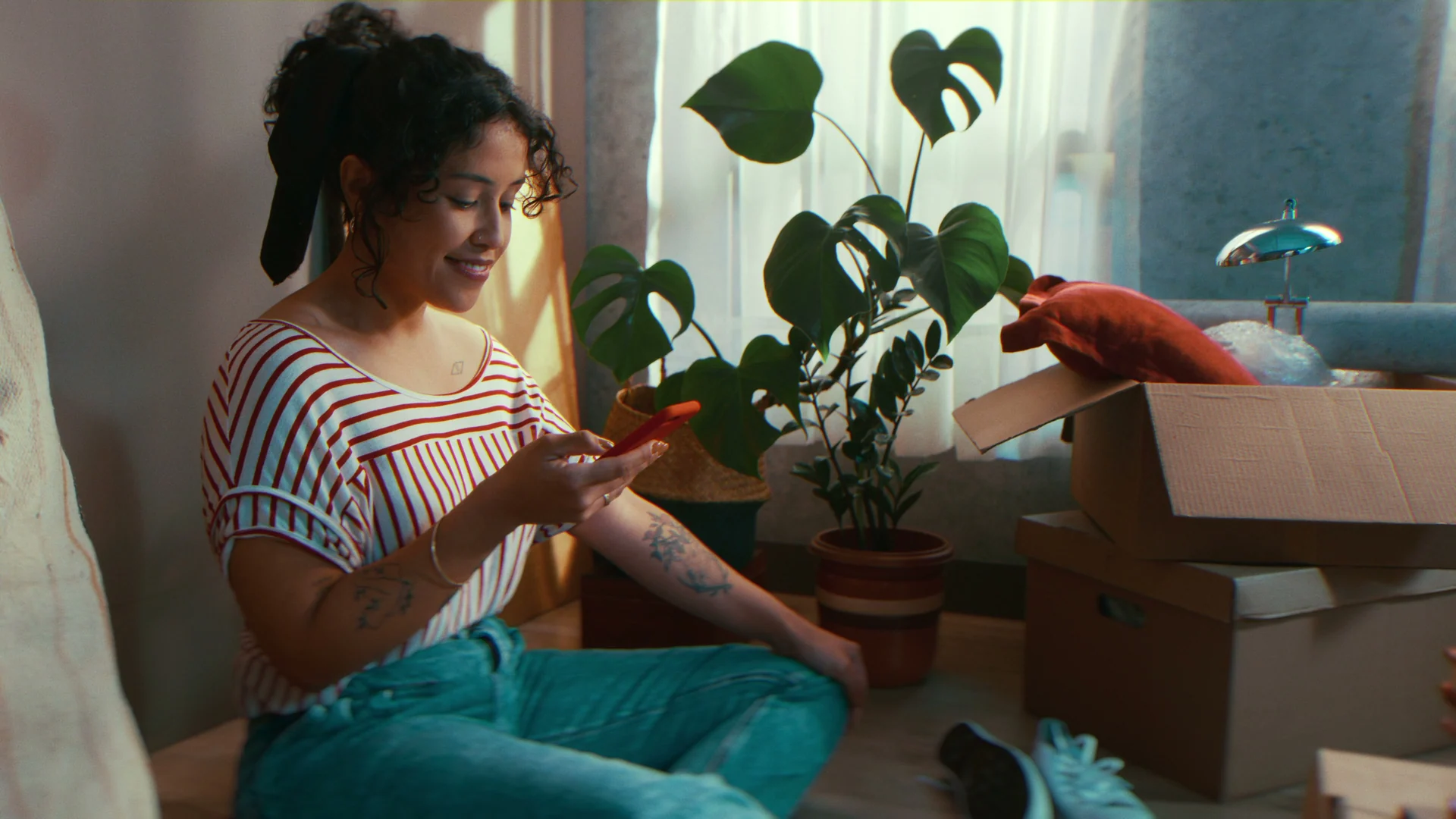 Mulher sorridente sentada no chão entre caixas de mudança e plantas, pesquisando móveis no celular