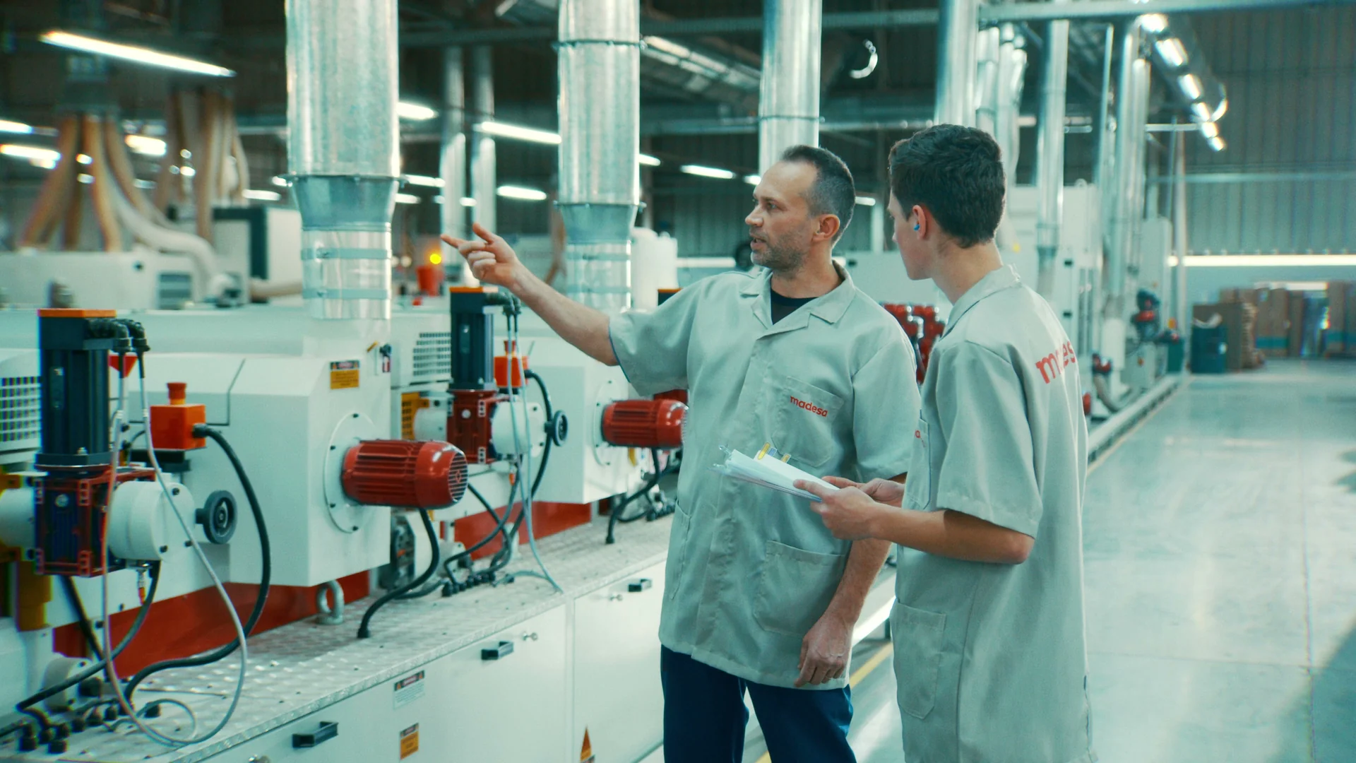 Dois operários de uniforme cinza conversando ao lado de máquinas da linha de produção
