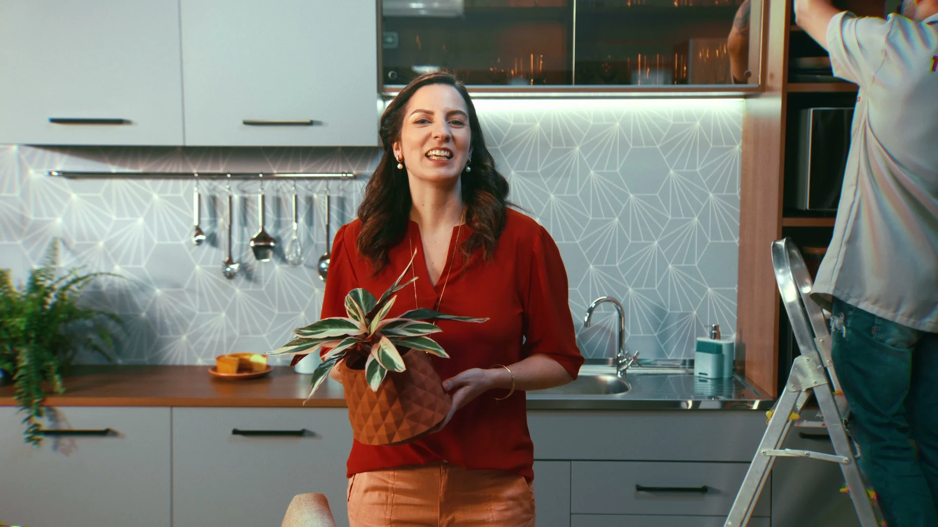 Mulher de blusa vermelha segurando um vaso de planta numa cozinha montada com armários brancos