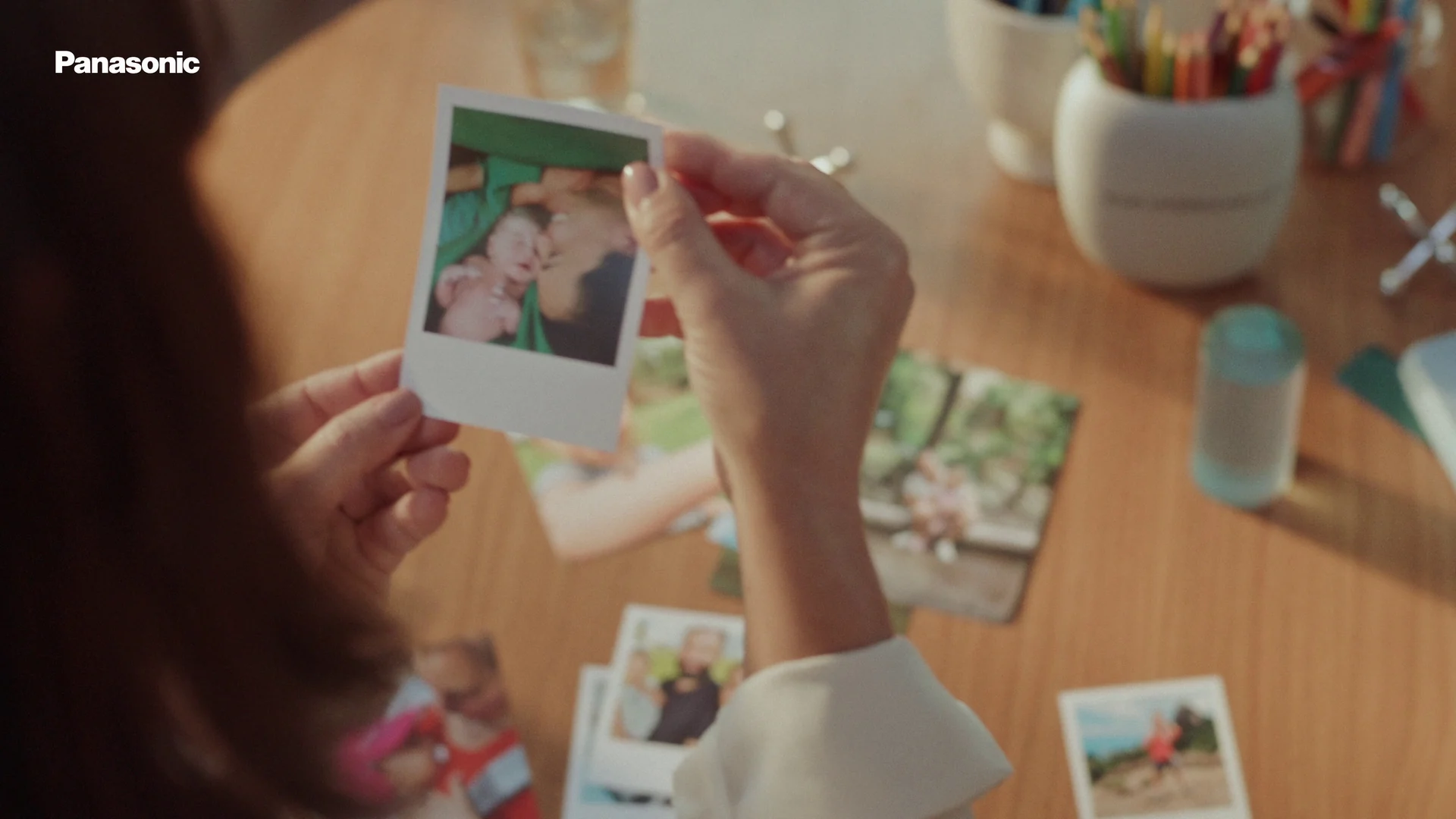 Mãos seguram uma foto instantânea de mãe e bebê sobre uma mesa de madeira cheia de outras fotos, lápis coloridos e um copo.