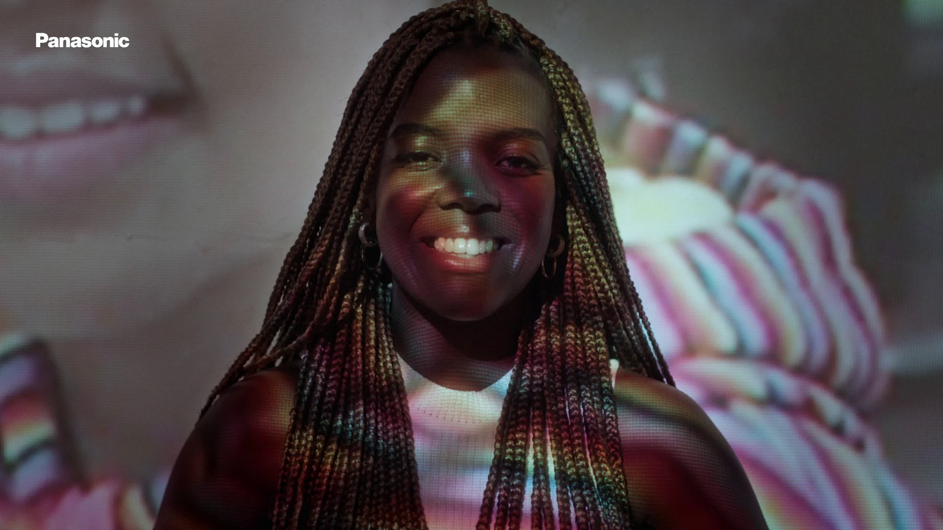 Retrato frontal de mulher de tranças sorrindo, com imagens coloridas projetadas sobre o rosto e a blusa.