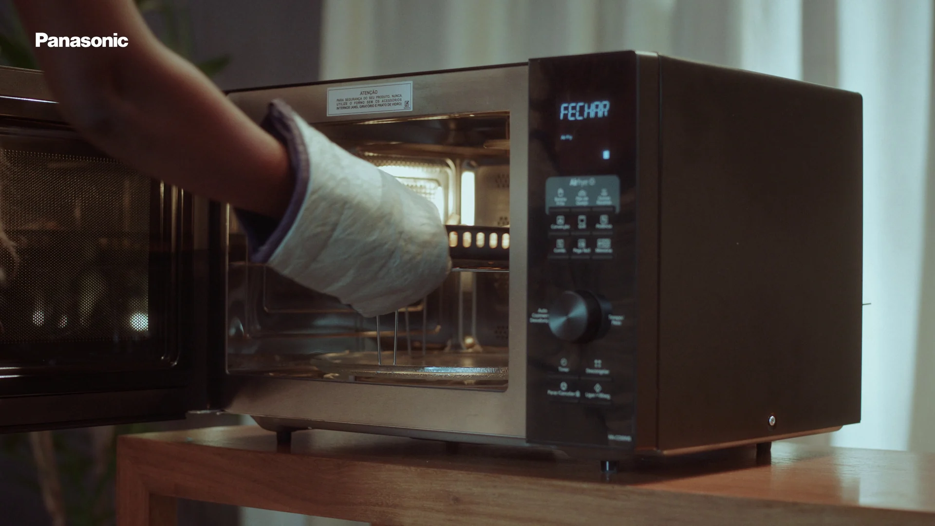 Mão com luva térmica retira uma bandeja de dentro de um micro-ondas Panasonic de inox sobre uma mesa de madeira.