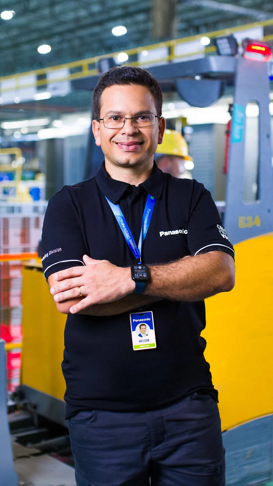 Colaborador de óculos, camisa polo preta Panasonic e crachá, de braços cruzados, com a linha de produção desfocada ao fundo.