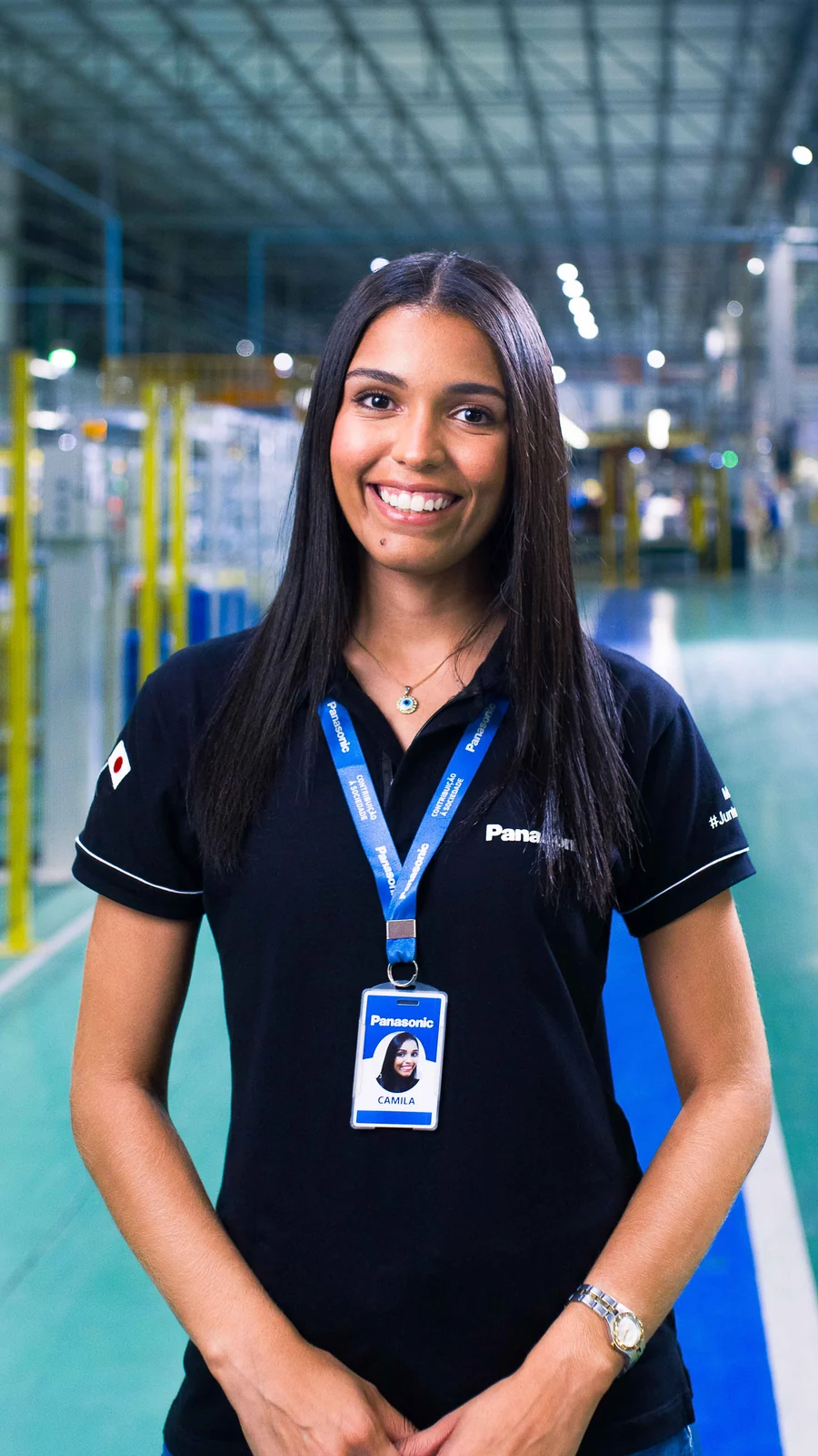 Colaboradora de camisa polo preta e crachá Panasonic sorri no corredor de piso azul e verde da fábrica.
