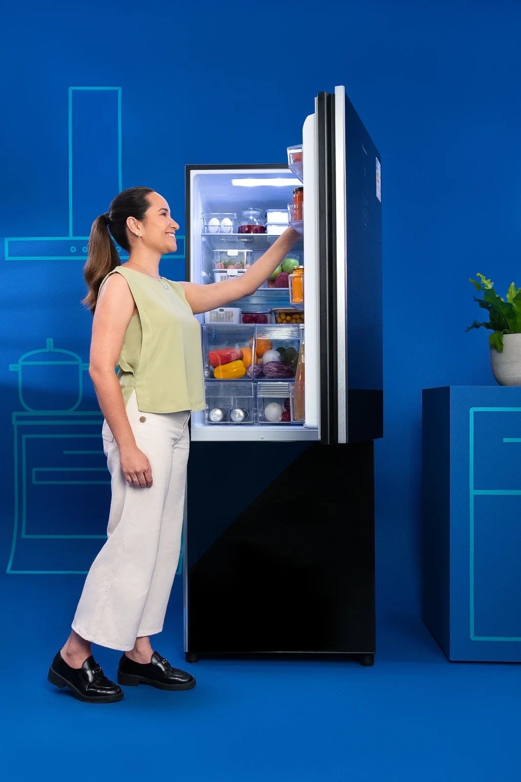 Mulher abre uma geladeira Panasonic de porta preta cheia de frutas e verduras, em cenário azul.