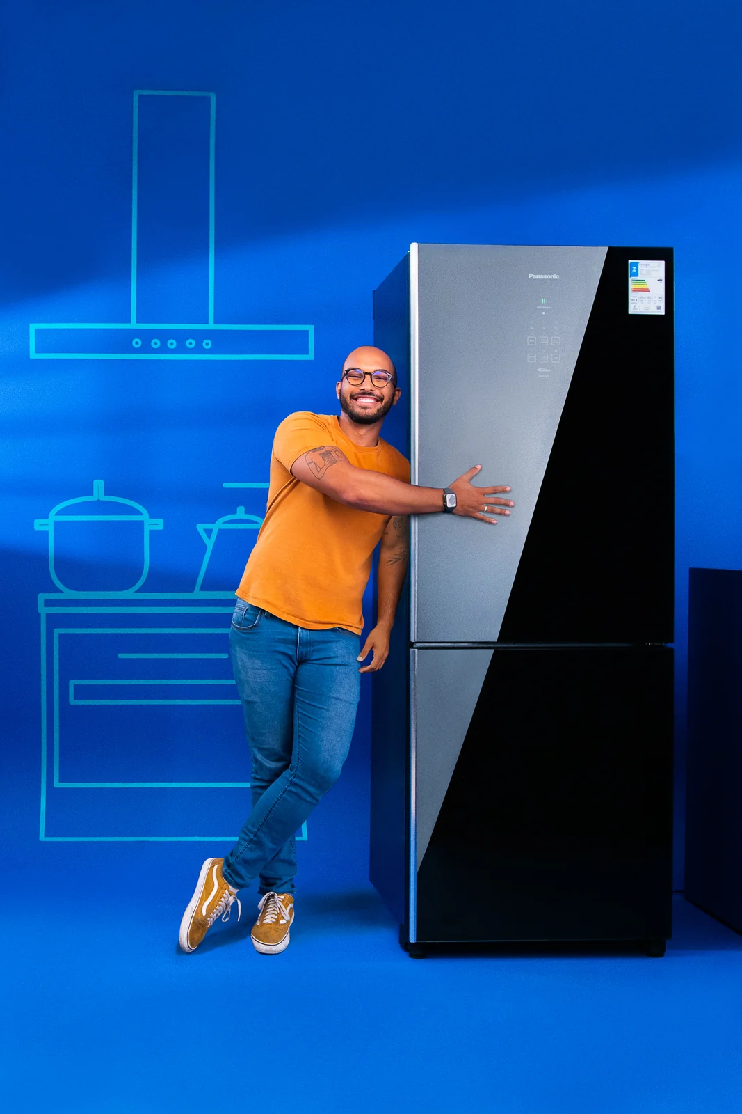 Homem de camiseta laranja abraça, sorrindo, uma geladeira Panasonic de porta espelhada no estúdio azul.