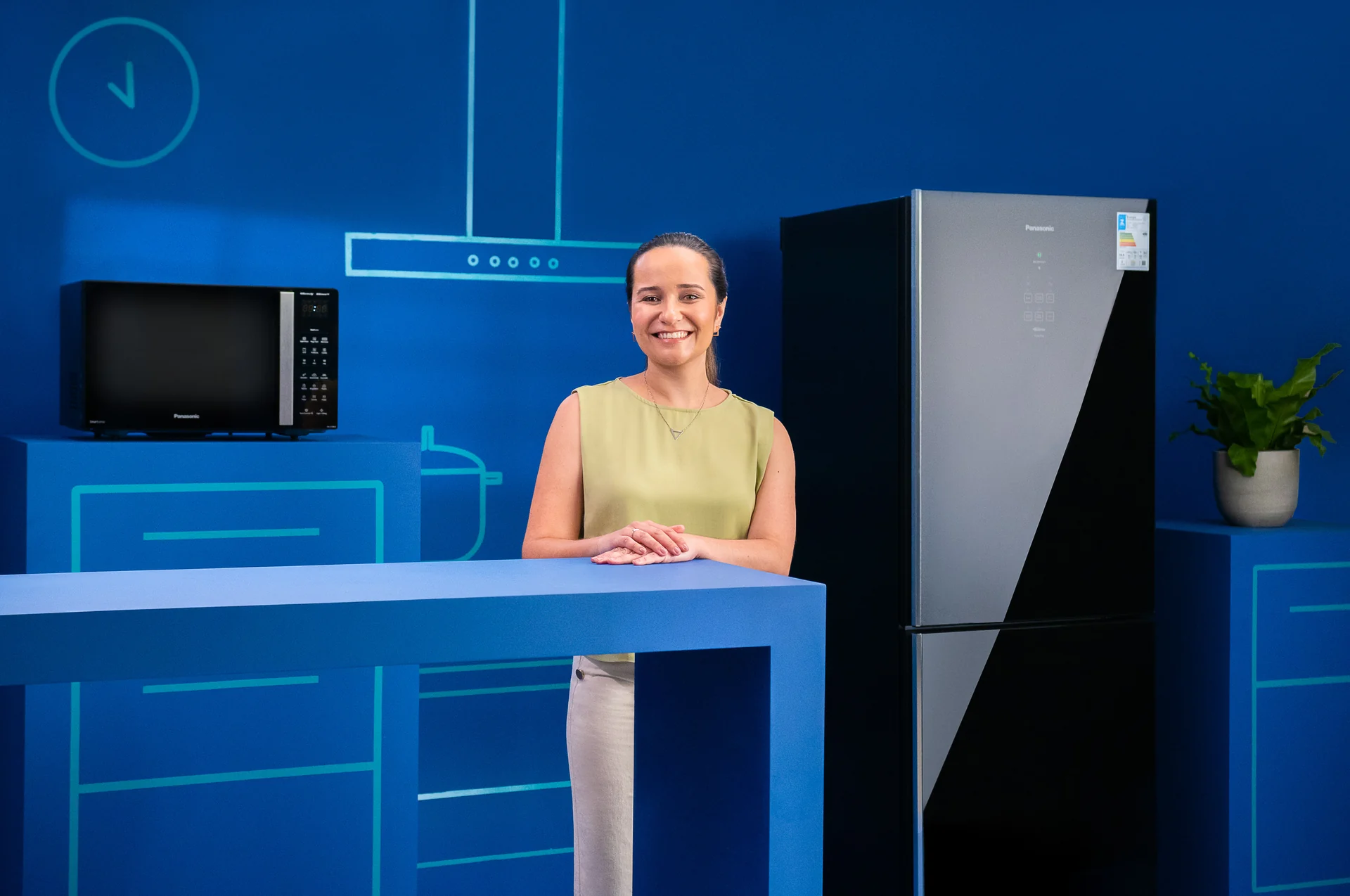 Colaboradora sorri atrás de uma bancada azul com micro-ondas, ao lado de uma geladeira Panasonic de porta espelhada, em cenário azul com grafismos de cozinha.