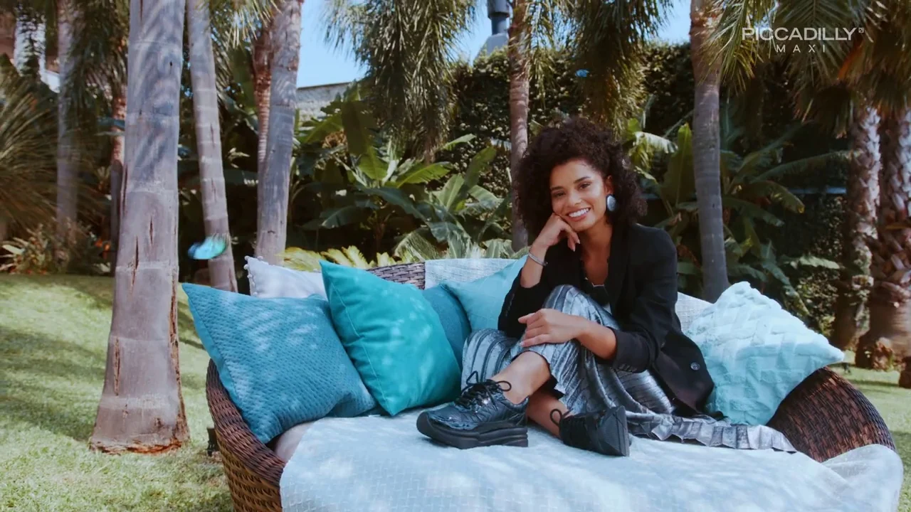 Modelo de blazer preto posa sentada em um sofá de vime com almofadas azuis, calçando tênis preto.