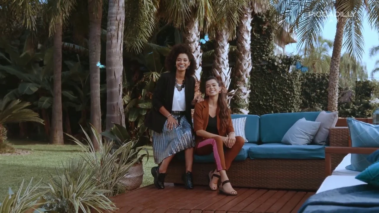 Duas modelos sorriem sentadas em um sofá azul-petróleo sob palmeiras, com borboletas azuis animadas ao redor.