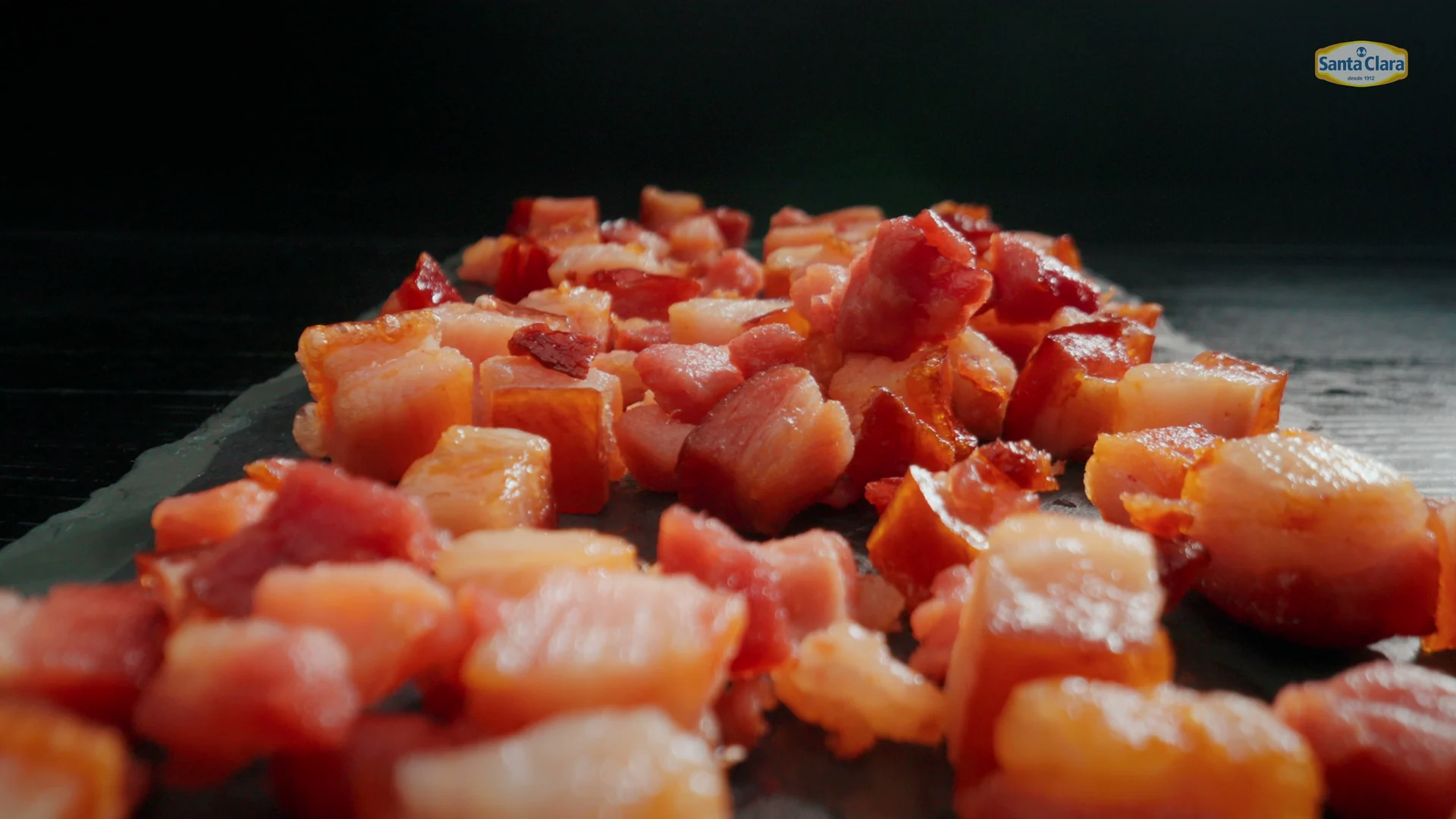 Cubos de bacon dourado empilhados sobre uma tábua de ardósia escura.