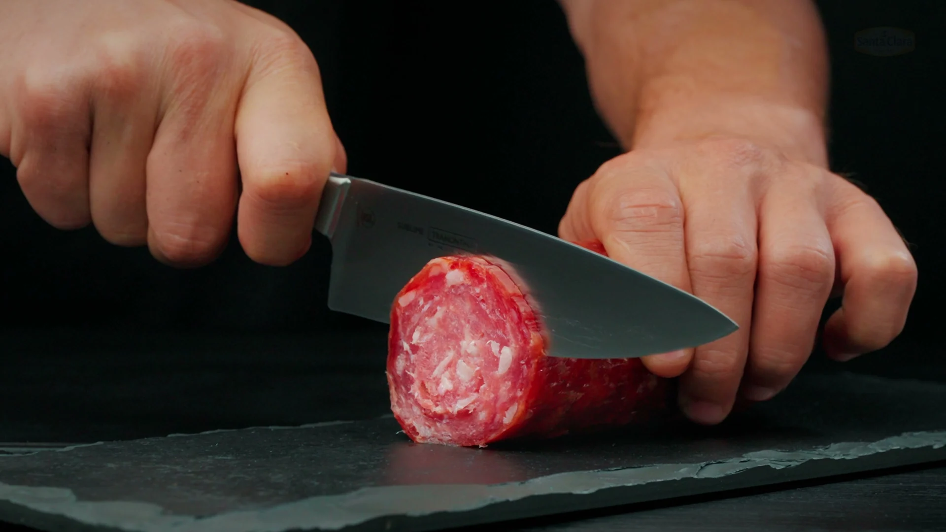 Mãos cortam um salame inteiro, mostrando o marmoreio da carne e da gordura.