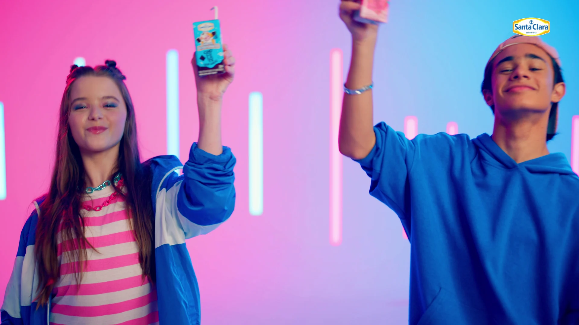 Menina e menino erguem caixinhas de bebida láctea Santa Clara diante de barras de neon rosa e azul.