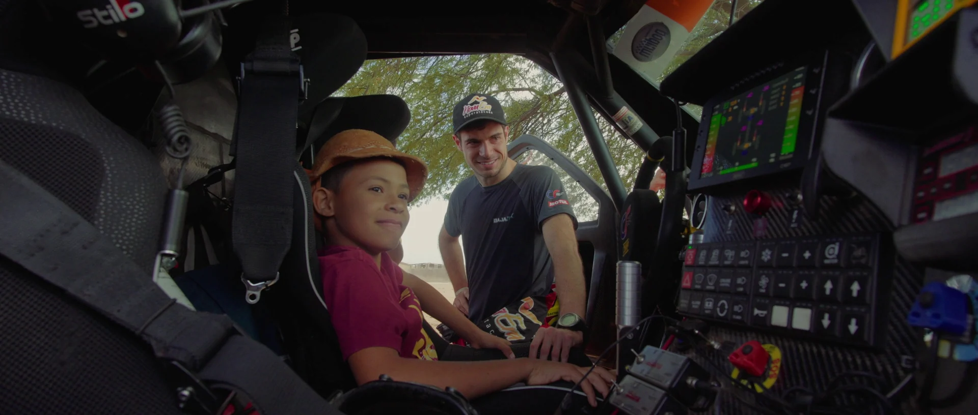 Menino de chapéu sentado no cockpit de um carro de rali conversa com um homem de boné do lado de fora.