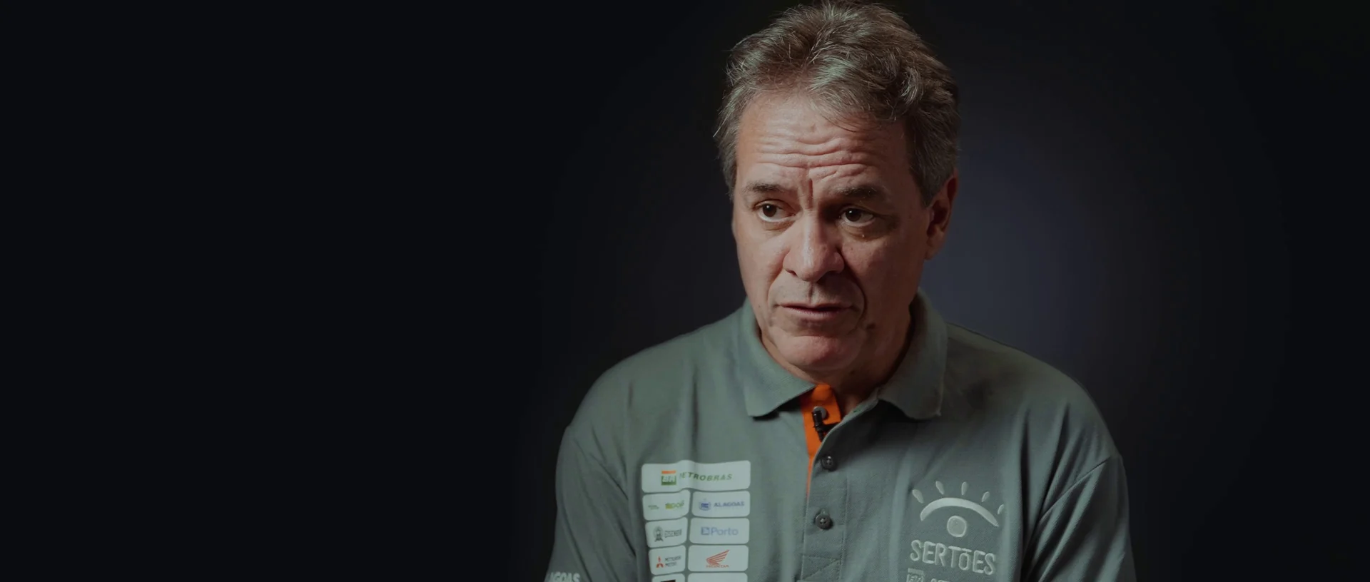 Homem grisalho de camisa polo do Sertões dá depoimento diante de fundo escuro.