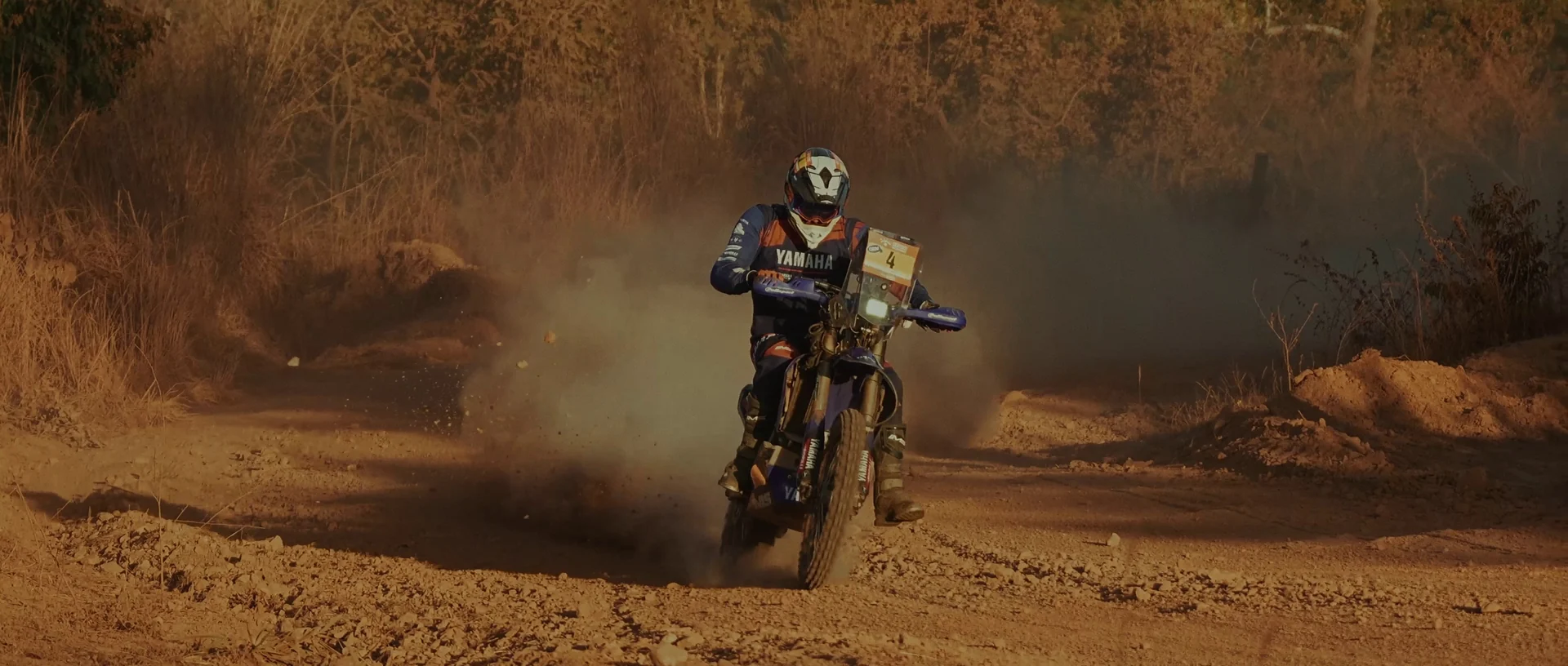 Piloto de moto atravessa uma curva de terra em meio à poeira e à vegetação seca.