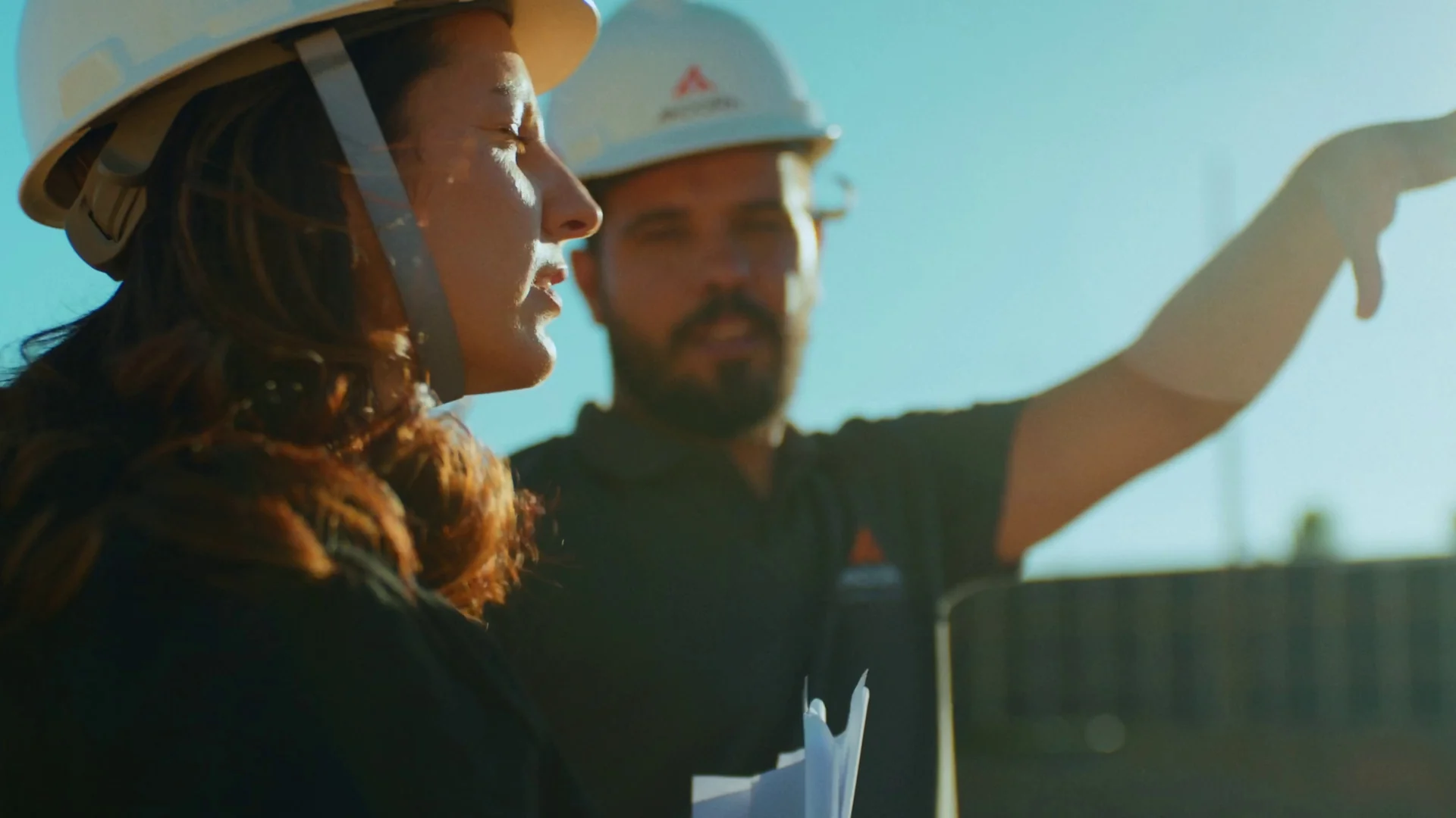 Mulher e homem de capacete branco observam uma obra sob céu azul; ele aponta para o alto.