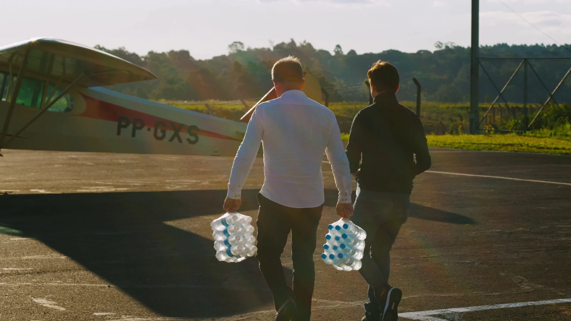 Dois homens caminham de costas por uma pista de pouso, carregando fardos de garrafas de água ao lado de um avião de asa alta, em luz de fim de tarde.