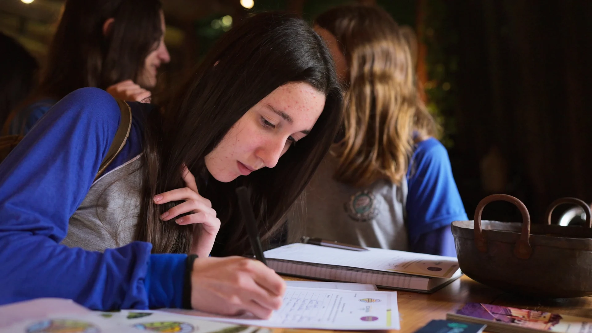 Estudante de uniforme azul escreve concentrada num caderno, com colegas desfocadas ao fundo.