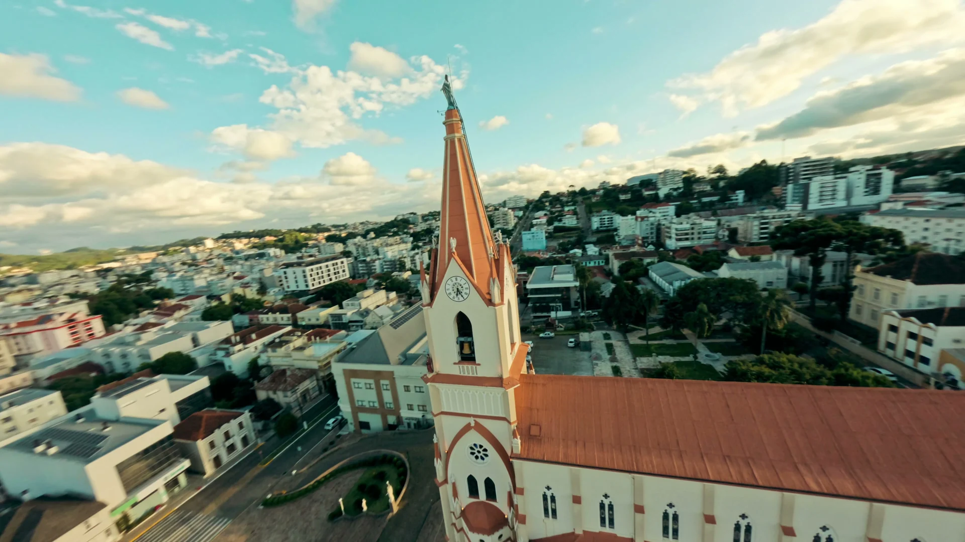 Voo baixo ao lado da torre de uma igreja de telhado vermelho, com a cidade e um céu de nuvens ao fundo.