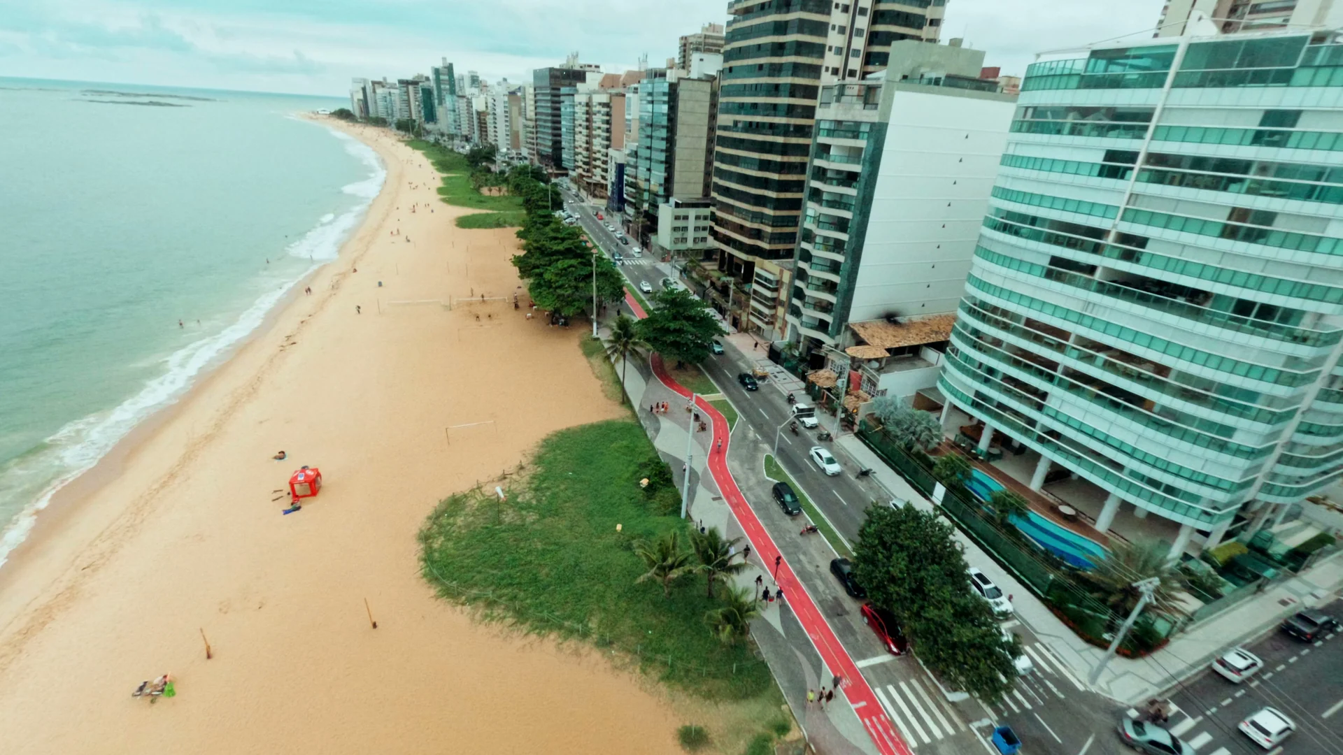 Orla vista do alto: praia de areia dourada, mar esverdeado e ciclovia vermelha entre edifícios.