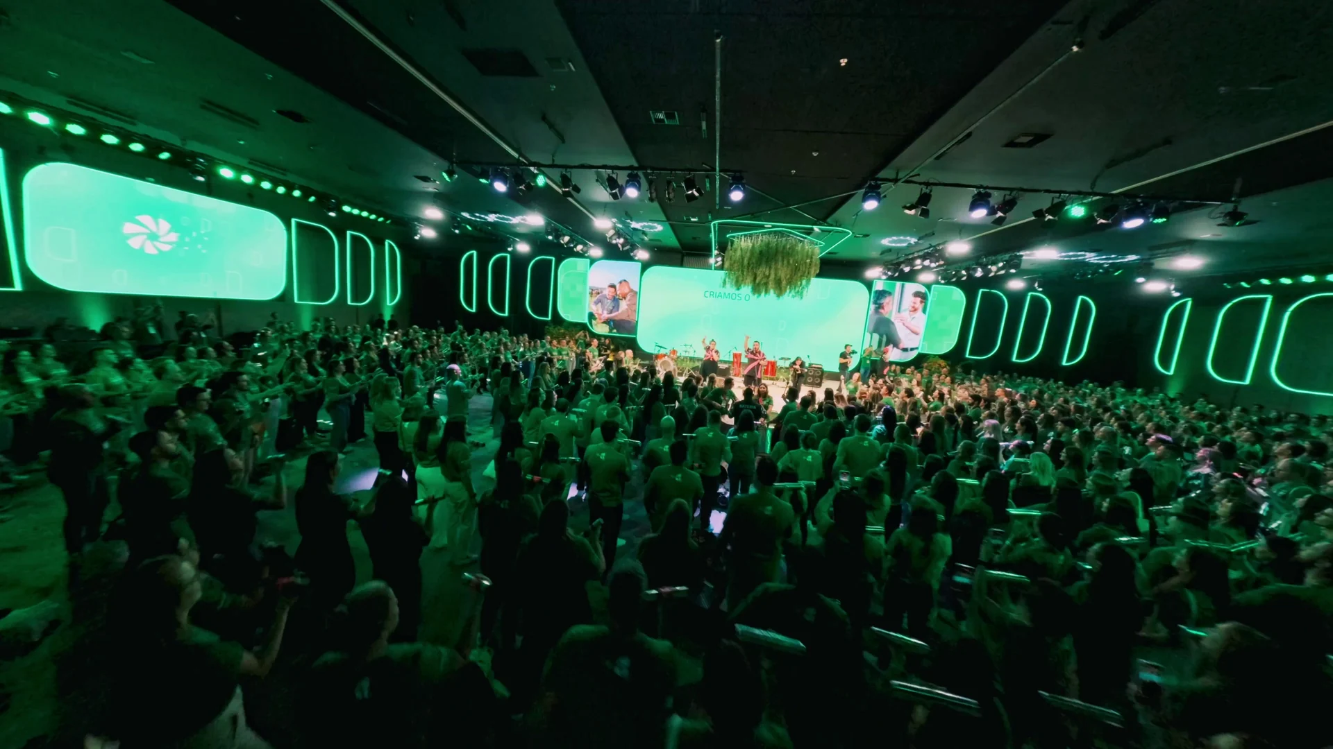 Auditório lotado de associados de camiseta verde diante de um palco com painéis de LED arredondados e iluminação verde.