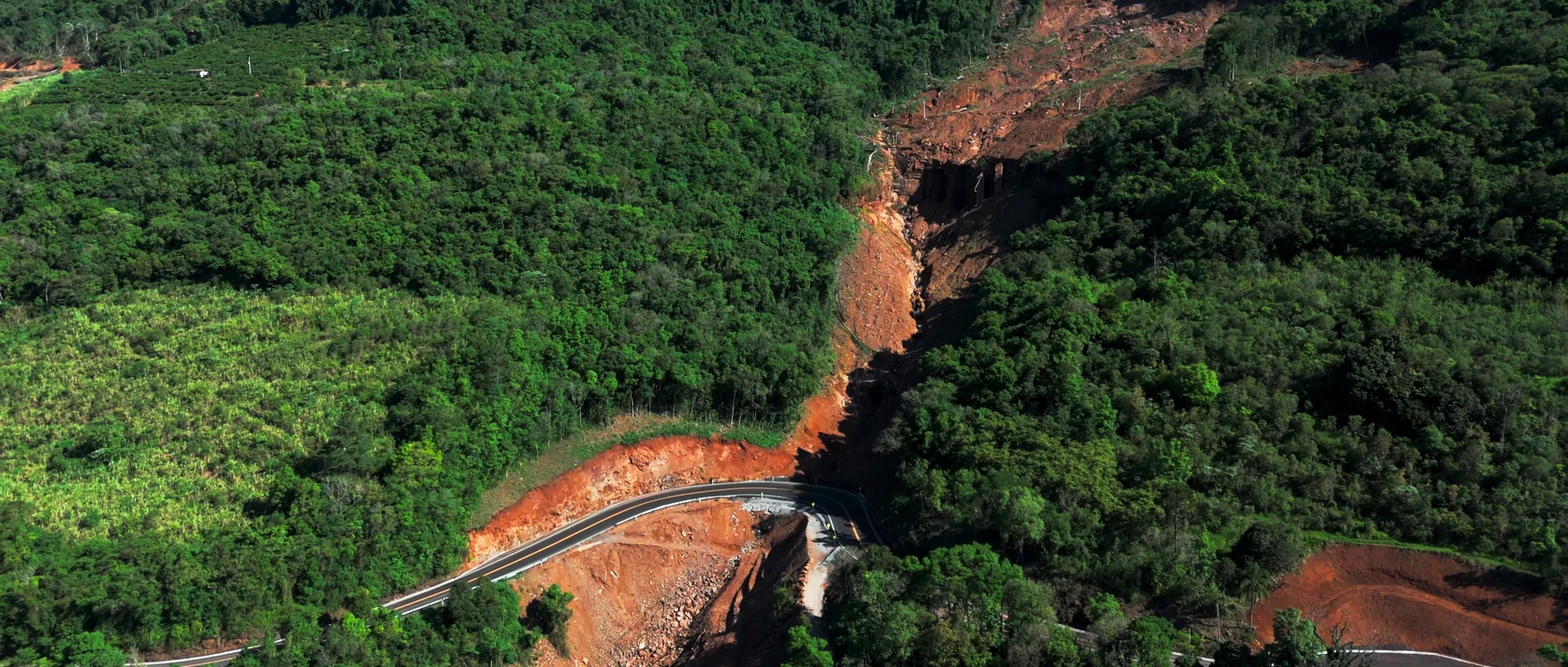 Vista aérea de uma encosta de mata cortada por um deslizamento de terra vermelha que atravessa uma estrada.