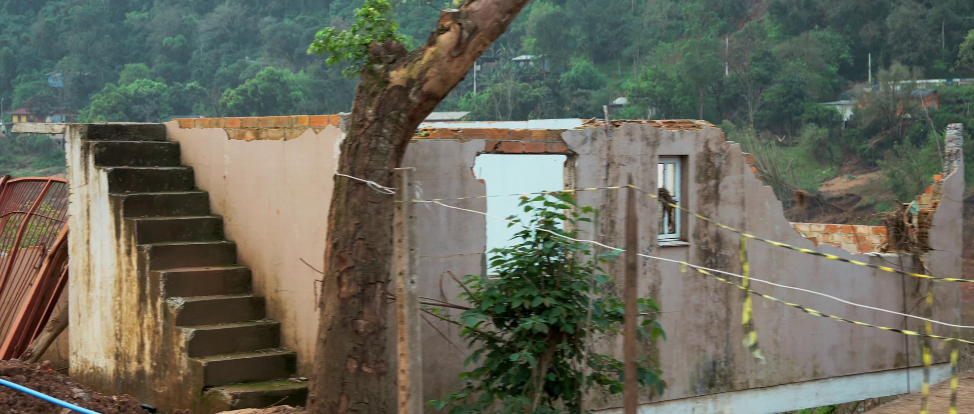 Ruína de uma casa sem telhado, com uma escada de concreto que termina no vazio e fita de isolamento amarela presa às paredes.